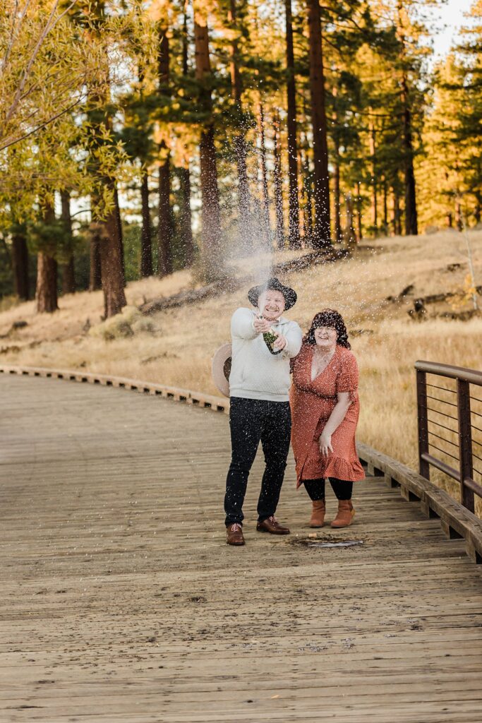 A couple is standing on a wooden walkway at Lake Tahoe with champagne in their hands.