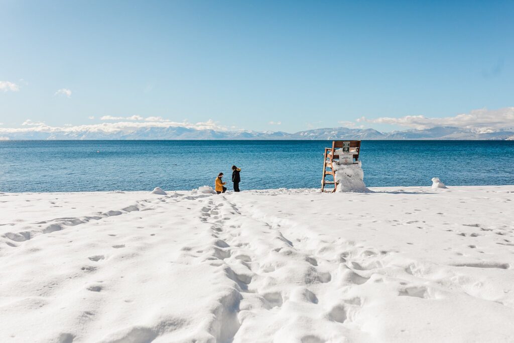 Two people standing on a snowy beach near lake tahoe.