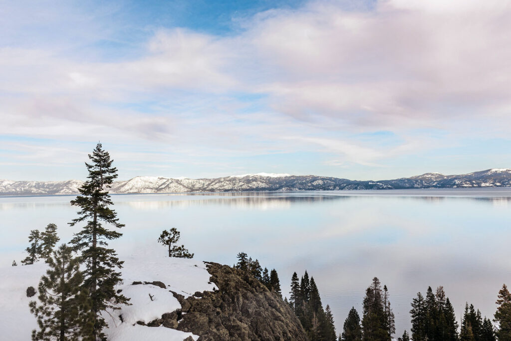 Discover Lake Tahoe's serene beauty with a calm, reflective lake surrounded by snow-covered mountains and pine trees under a partly cloudy sky, offering a perfect escape from the hustle and bustle.