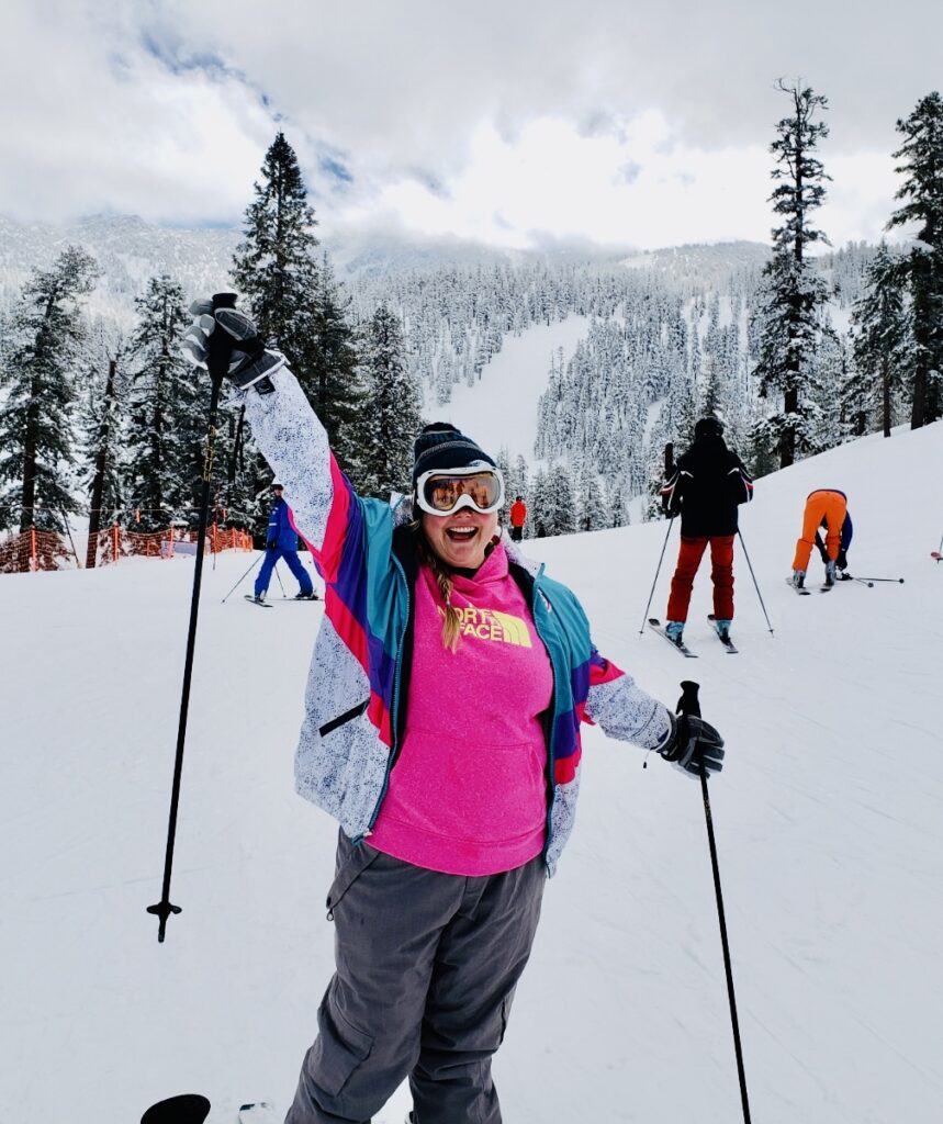 A person in a colorful ski outfit stands joyfully on a snowy Lake Tahoe slope with ski poles raised, surrounded by other skiers and snow-covered trees under a cloudy sky—a perfect moment to discover Lake Tahoe.