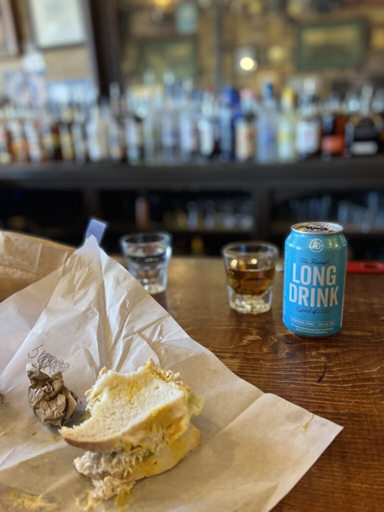 A half-eaten sandwich on paper sits next to an unopened can of Long Drink and two glasses on a bar counter, echoing the casual vibe one might discover in Lake Tahoe's lively après-ski scene, with a row of bottles lining the background.