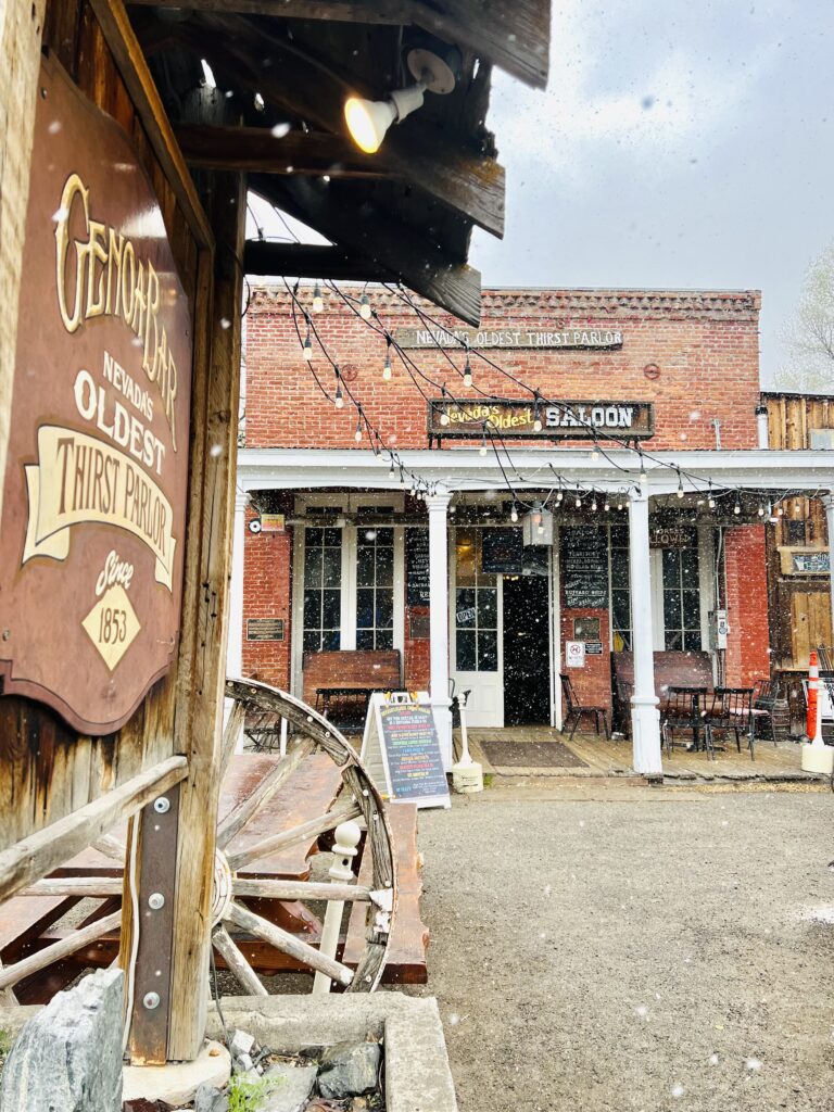 Exterior of a rustic brick saloon with a sign reading "Nevada's Oldest Thirst Parlor." A covered porch and chalkboard menu are visible, with light snow falling. As you explore things to do in Lake Tahoe, be sure to stop by and experience its historic charm.