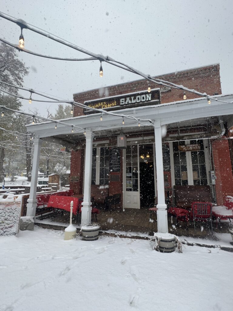 A red brick saloon with a snow-dusted porch and string lights, perfect for those looking to discover Lake Tahoe. Snow covers the ground and benches outside, making it a cozy spot on your list of things to do in Lake Tahoe.