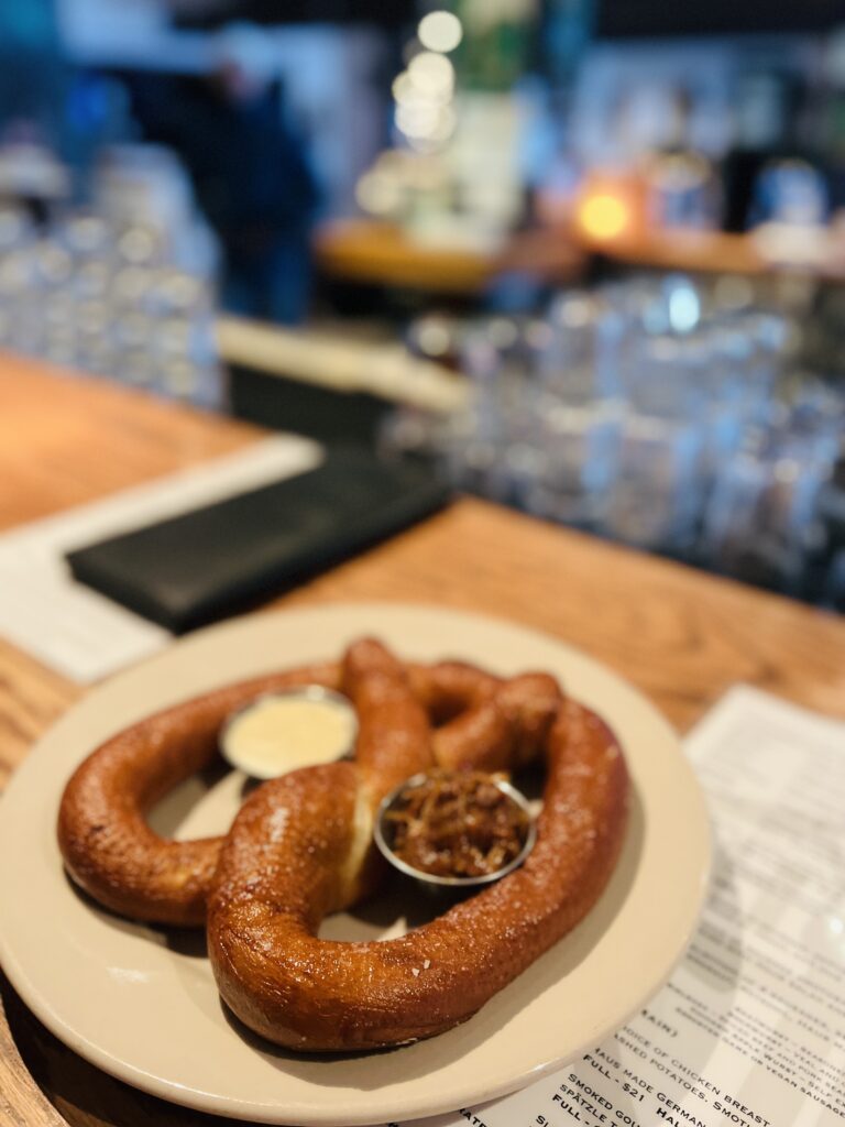 A large pretzel with two sides of dipping sauce on a plate sits on a wooden table, reminiscent of the cozy après-ski treats you might enjoy while exploring things to do in Lake Tahoe, with a beautifully blurred background adding to the ambiance.