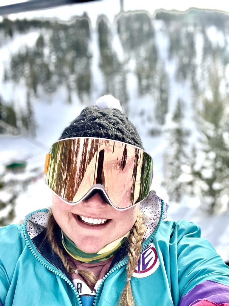 A person wearing ski goggles and a winter jacket smiles while on a ski lift, surrounded by snowy trees. Discover Lake Tahoe's stunning winter wonderland—a perfect start to exploring the many things to do in Lake Tahoe.