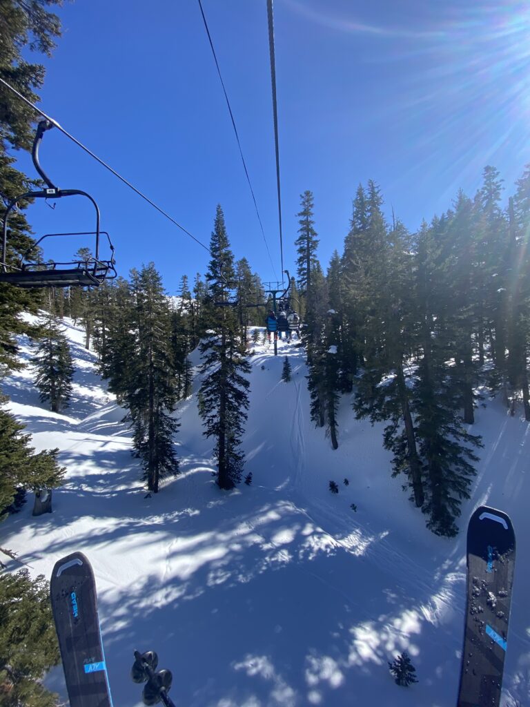 Discover Lake Tahoe from a ski lift: soak in the panoramic views of snow-covered slopes and towering pine trees under a clear blue sky. Skis peek into the frame, hinting at adventures that await in this winter wonderland.