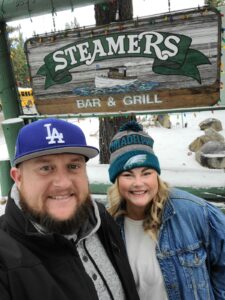 Two people, bundled in casual winter clothing and caps, are smiling in front of a "Steamers Bar & Grill" sign. Snow blankets the ground, adding to the charm of their adventure as they discover Lake Tahoe's delightful spots.