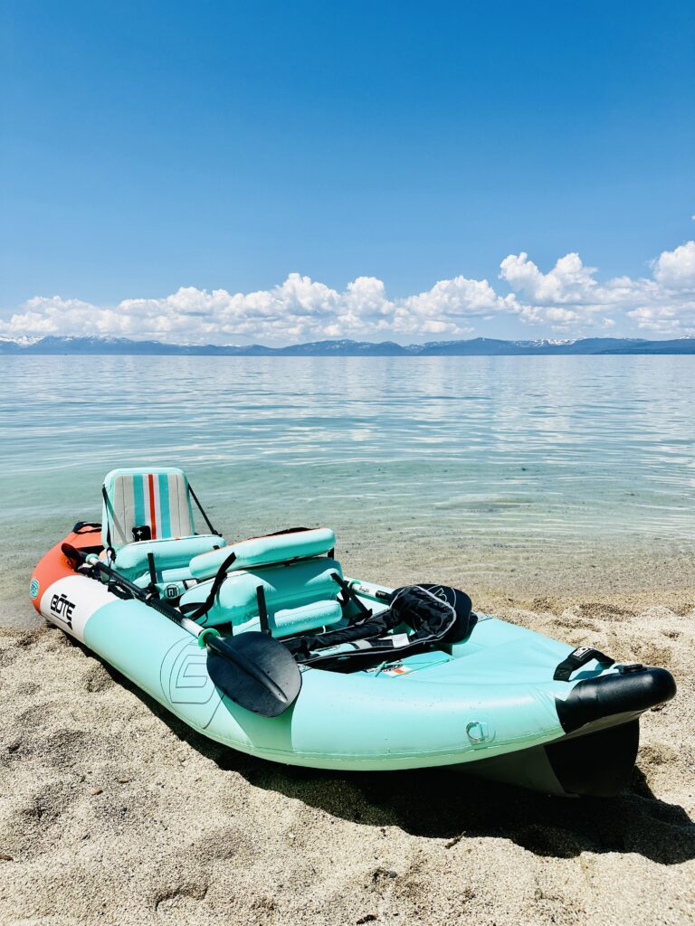 A teal inflatable kayak with paddles rests on a sandy beach by a calm lake under a clear blue sky—perfect for those looking to discover Lake Tahoe and add to their list of things to do in Lake Tahoe.