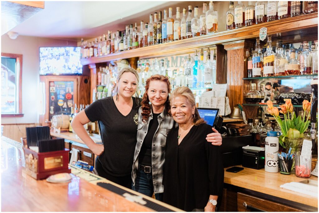 Three women stand behind a bar counter filled with bottles, smiling at the camera, embodying the welcoming spirit you can discover in Lake Tahoe. The middle woman has an arm around each of the others, ready to share tips on things to do in Lake Tahoe.