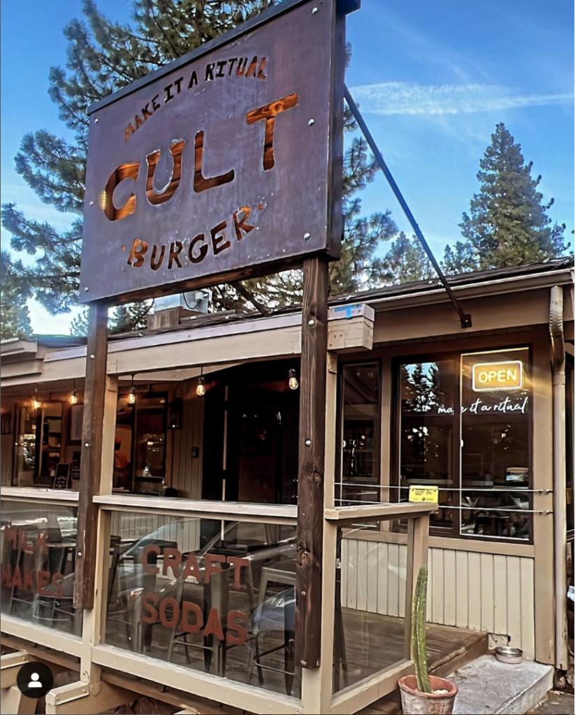 Exterior view of a burger restaurant with a sign reading "Cult Burger" and "Make it a Ritual." A neon "Open" sign glows in the window. There's outdoor seating beside a cactus, making it the perfect pit stop on your journey to discover Lake Tahoe.