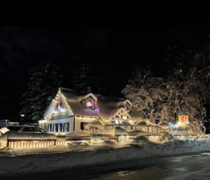 A snowy house at night with warm, glowing lights along the roof and fence invites you to discover Lake Tahoe. A small sign with an emblem is visible near the entrance, promising a cozy retreat amidst this winter wonderland.