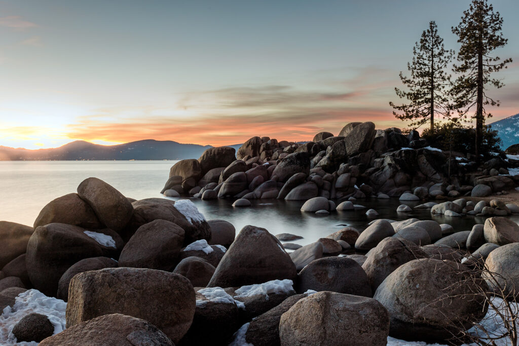 Discover Lake Tahoe's serene beauty on a rocky shoreline with boulders, two tall trees, and light snow. Enjoy the calm water and distant hills under a colorful sunset sky, making it one of the must-experience things to do in Lake Tahoe.