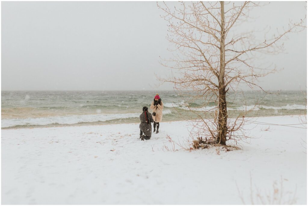 Two people, bundled in winter clothing, stand on a snow-covered beach by a tree, with ocean waves in the background.