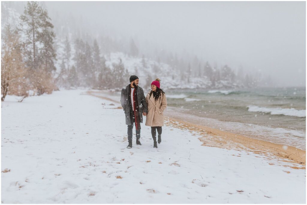 A couple walks hand in hand on a snow-covered beach with a lake and pine trees in the background. Snow is gently falling.