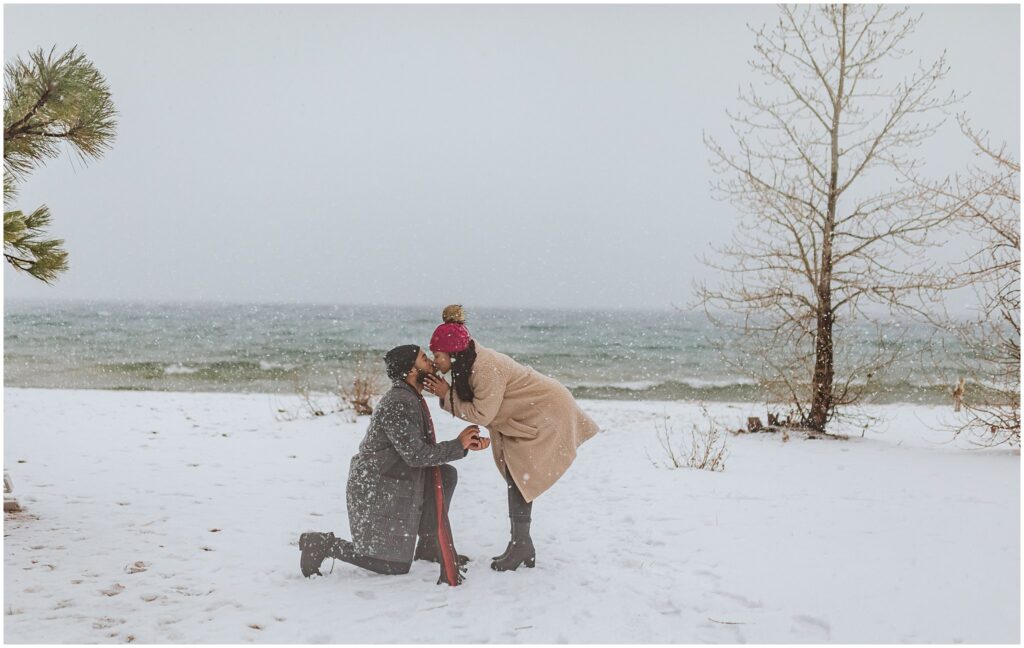 A person kneels in the snow proposing to another person by a snowy lakeside. Trees and the lake are visible in the background.