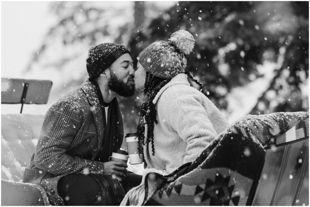 A couple shares a kiss in the snow, both wearing winter attire and holding coffee cups.