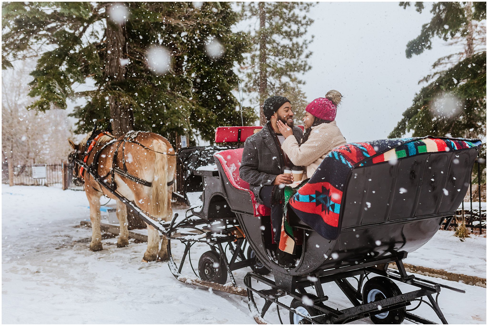 A couple sits in a horse-drawn sleigh surrounded by snow, under a colorful blanket. They're cozy, laughing, and drinking from mugs while snowflakes fall around them.