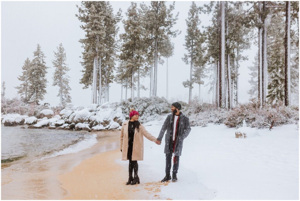 Two people wearing winter clothing hold hands and walk on a snowy beach surrounded by trees.