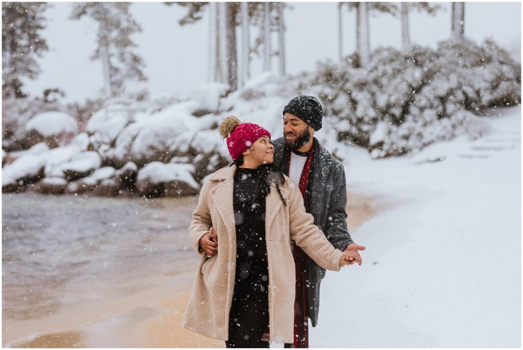 A couple stands on a snowy beach, smiling at each other. Snow is falling, and trees and rocks are in the background.