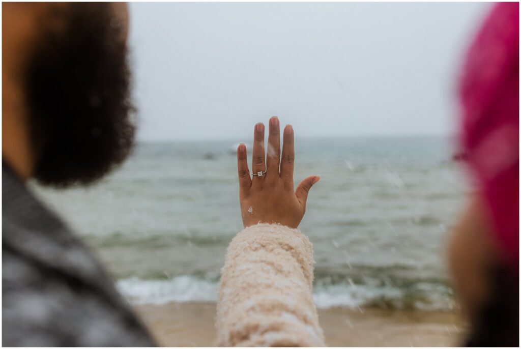 A person holds up their hand with a ring on their finger, facing the ocean. A second person is partially visible, wearing a pink hat.