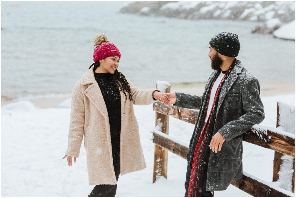 A woman and a man in winter clothing stand in the snow by a wooden fence, smiling at each other near a snowy lake.