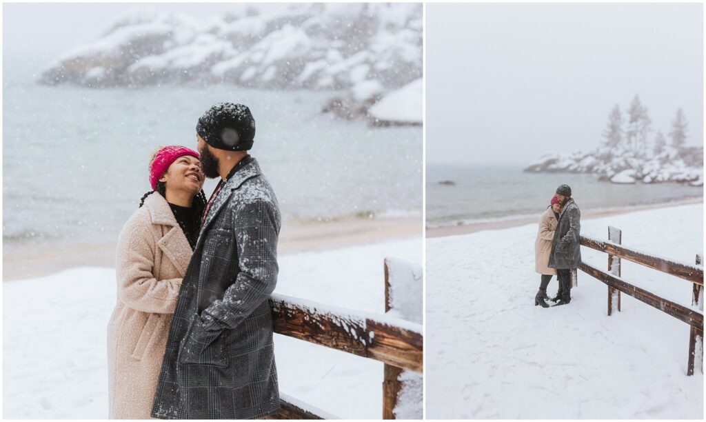 A couple stands by a snow-covered railing near a lake, embracing and smiling in a snowy landscape.