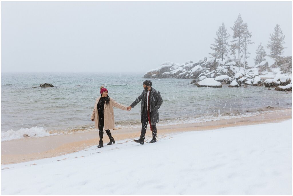 A couple walks hand in hand along a snowy beach, with overcast skies and rocky trees in the background.