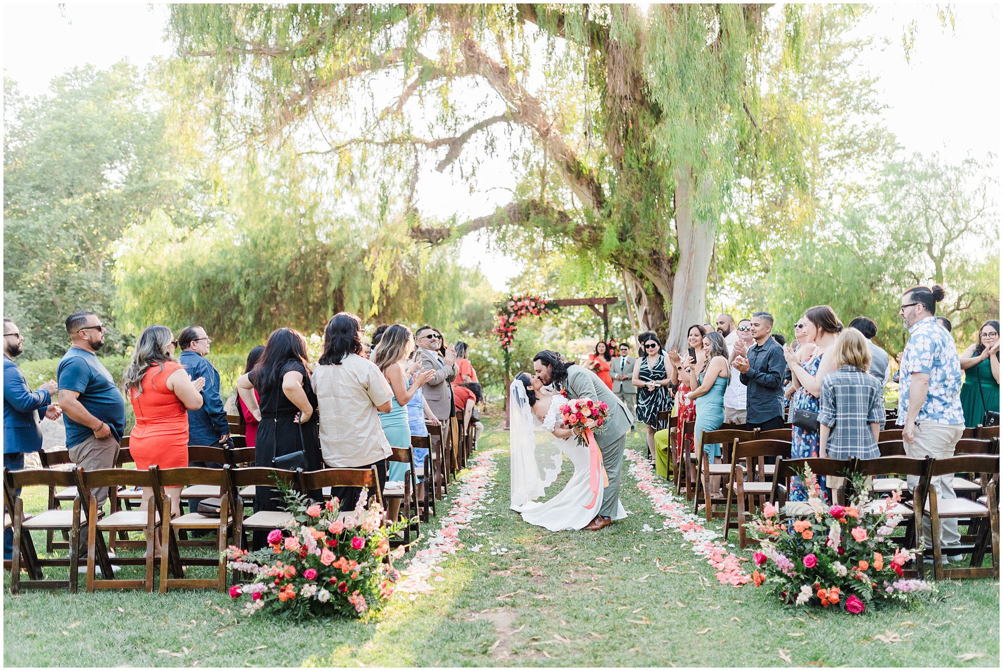 Bride and groom walking down an outdoor aisle, surrounded by seated and standing guests, with flowers lining the path and a large tree in the background.