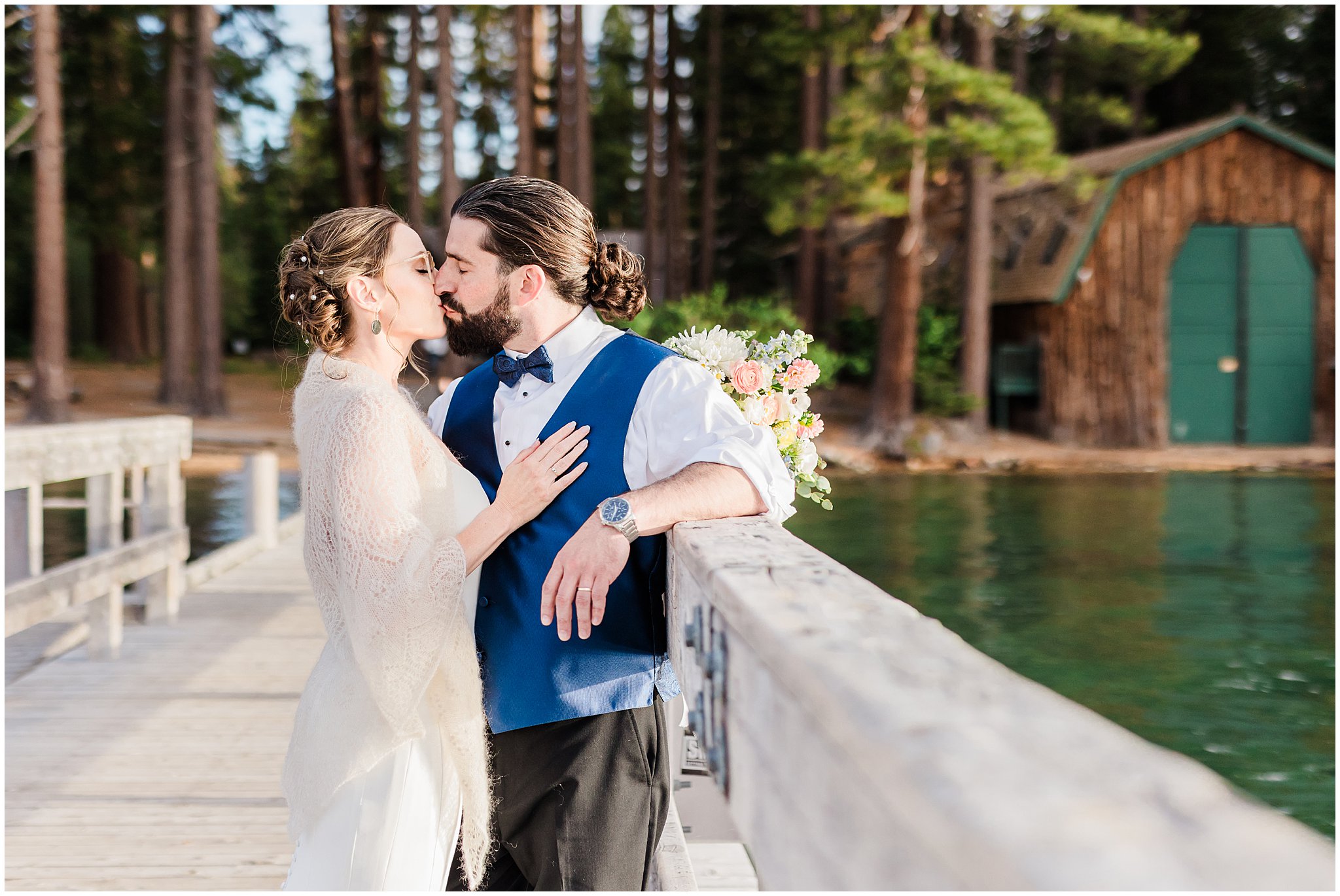 A couple kissing on a pier, with a lake and a wooden cabin surrounded by trees in the background. The man is wearing a blue vest and bow tie, and the woman is in a shawl with flowers.