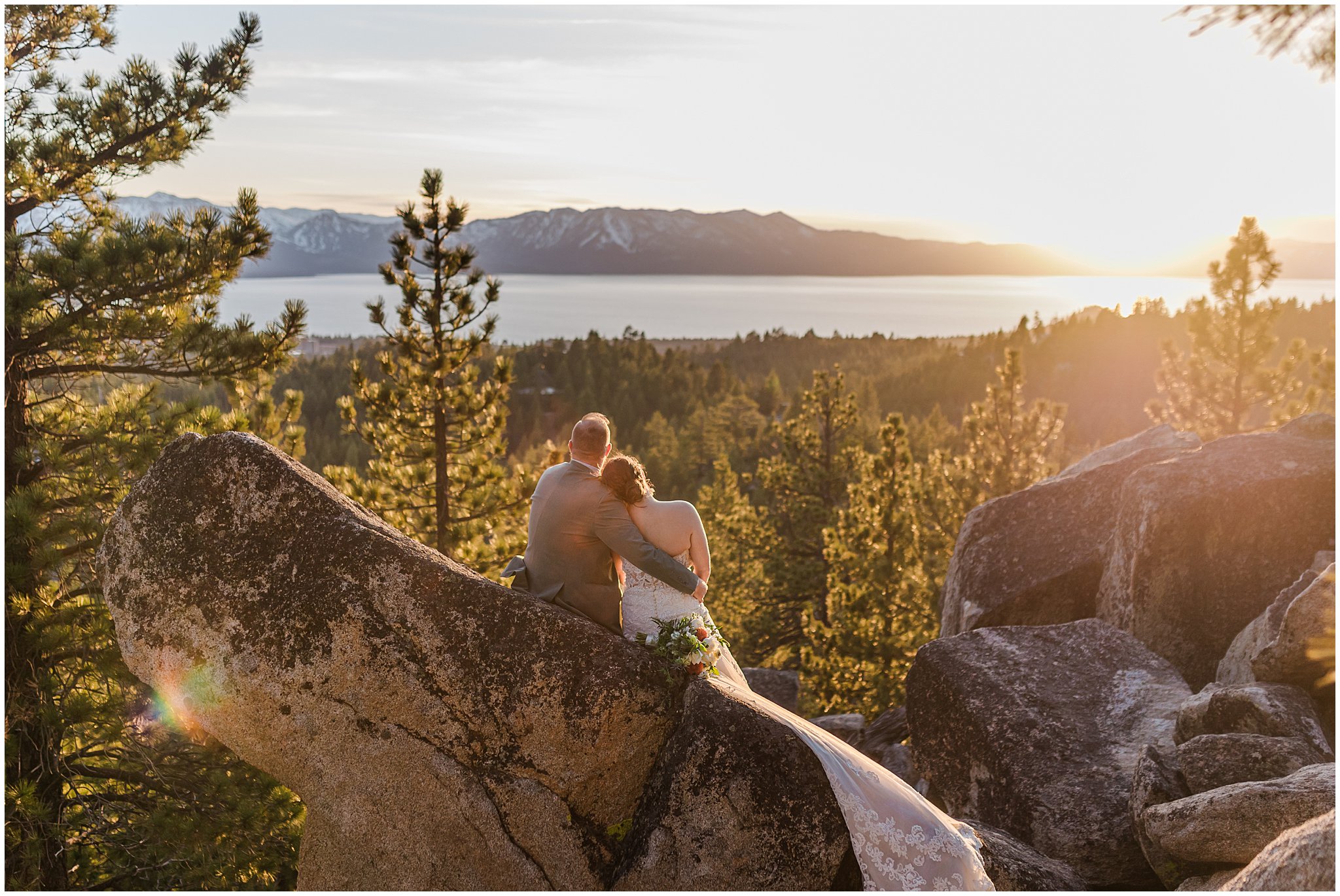 A couple in wedding attire sits on large rocks overlooking a scenic landscape with mountains, forest, and a lake at sunset.