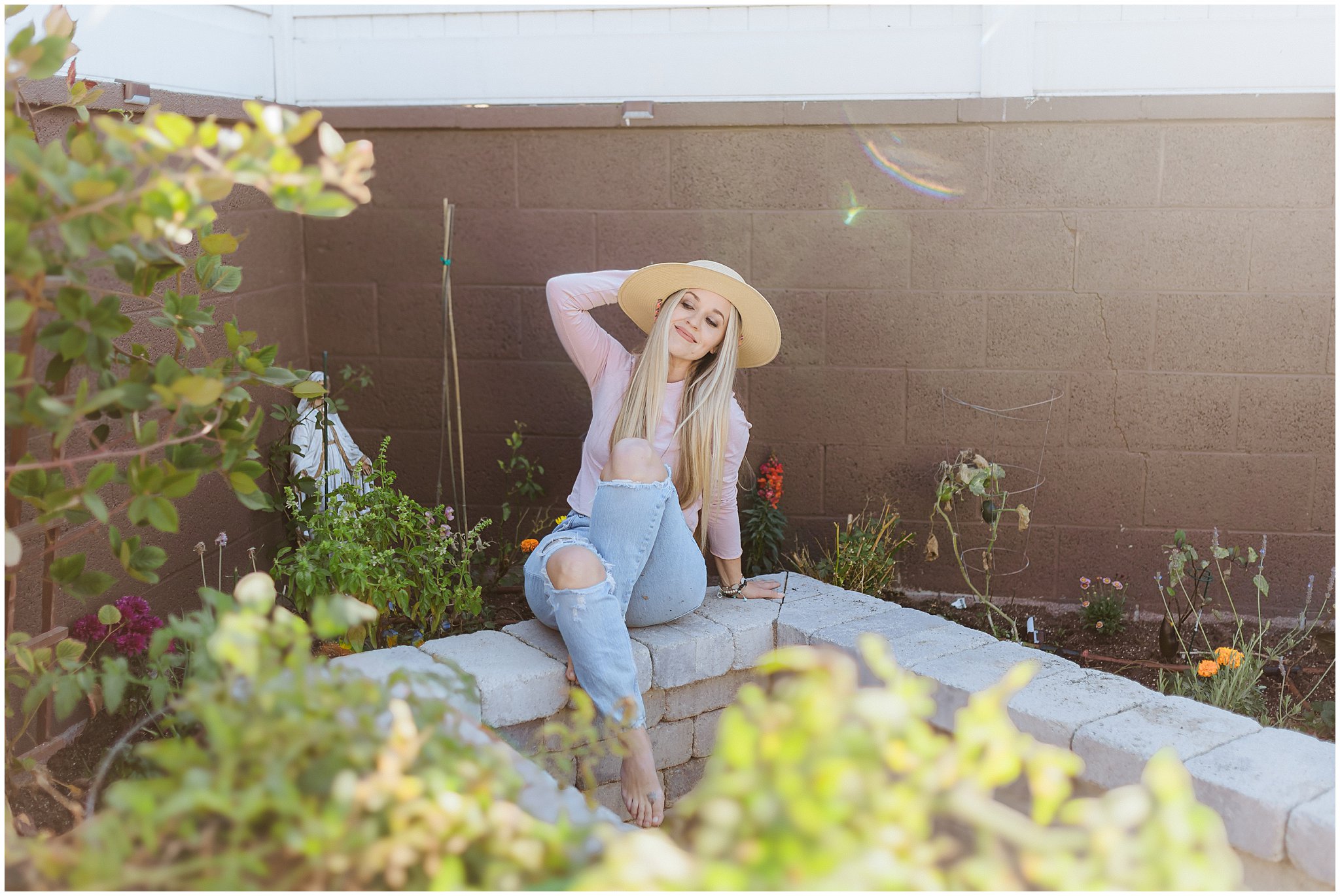 Person sitting on a stone ledge in a garden, wearing a straw hat and casual clothing. They appear relaxed, with one hand behind their head.