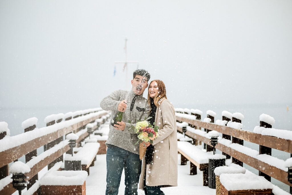A man pops a champagne bottle while standing next to a smiling woman holding flowers on a snowy pier during a magical Lake Tahoe winter proposal.