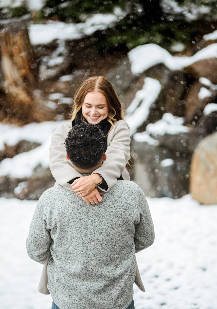 A woman smiling with her arms around a man's shoulders, standing together in a snowy outdoor setting during their Lake Tahoe winter proposal.
