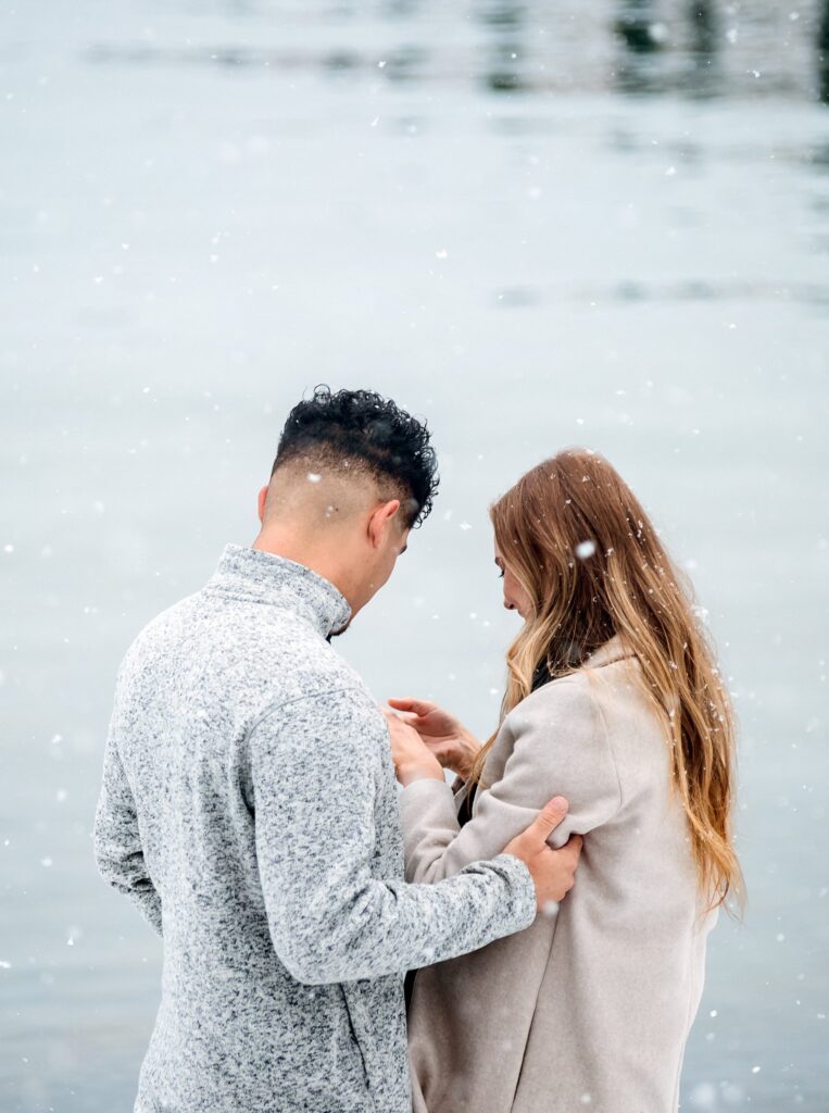 A man and woman stand close together by Lake Tahoe in light snowfall, facing each other and holding hands during a magical winter proposal.