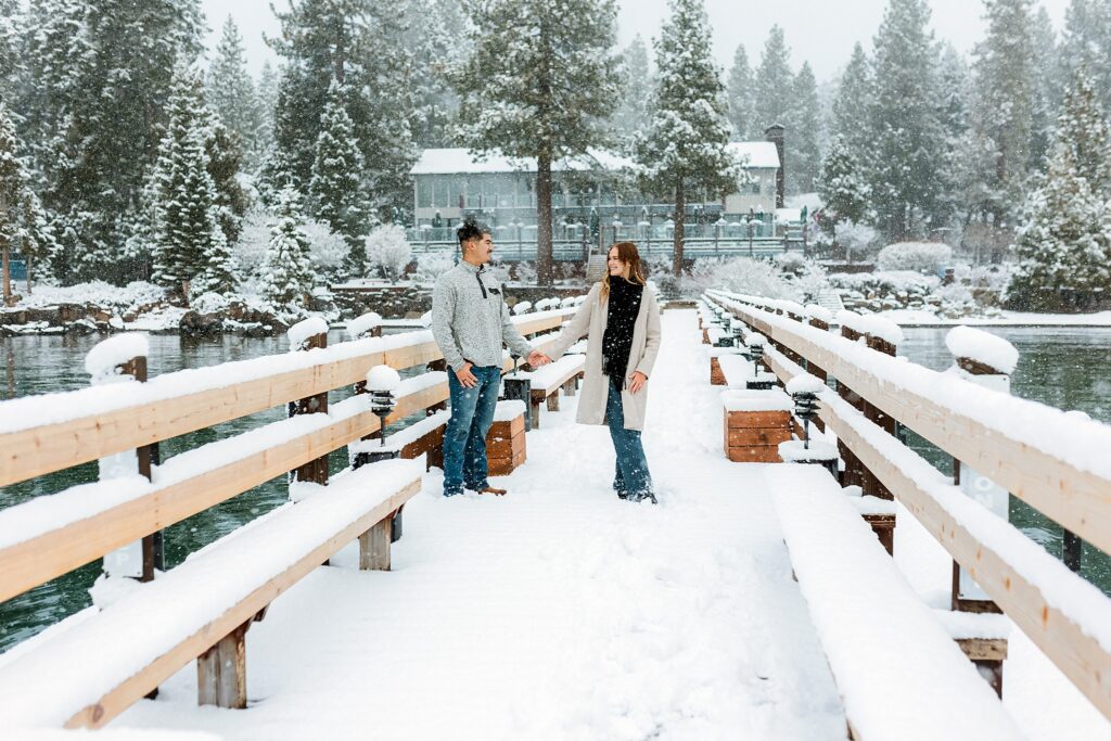 A man and woman stand holding hands on a snow-covered wooden bridge, the magic of a Lake Tahoe Winter Proposal reflected in the icy lake and snow-draped trees behind them.