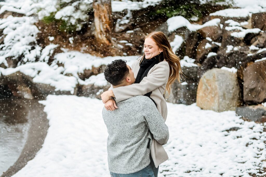 A man lifts a woman off the ground as they smile at each other outdoors, surrounded by snow-covered rocks and trees during a magical Lake Tahoe Winter Proposal.