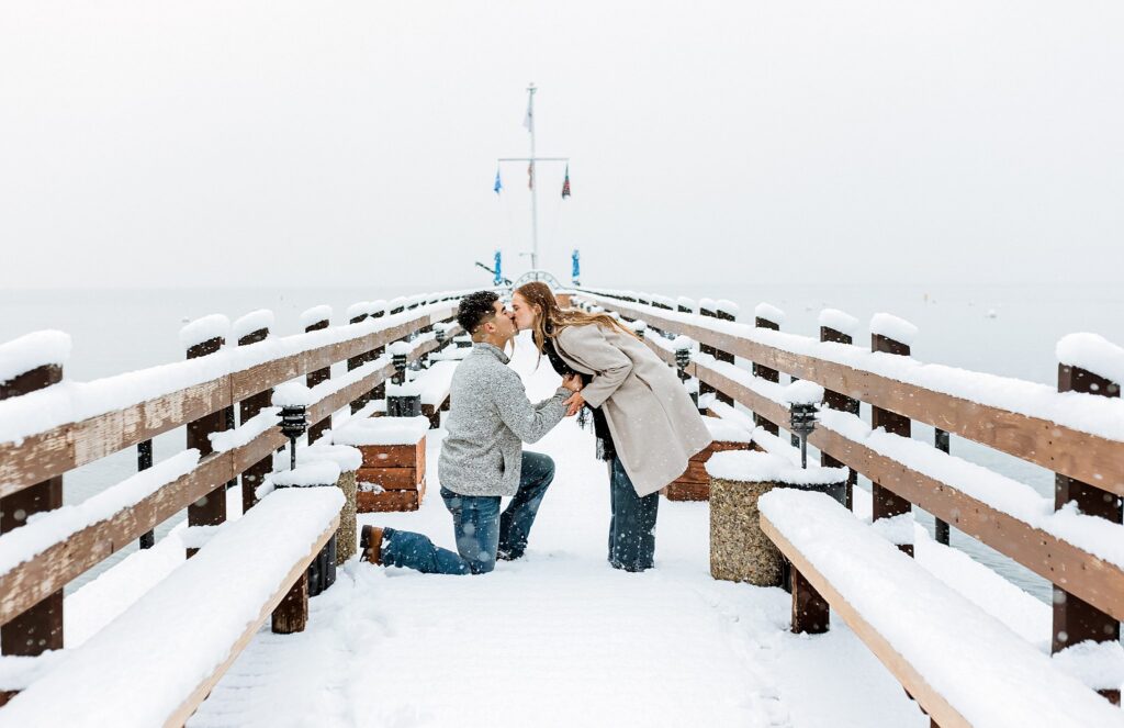 A man kneels and proposes to a woman on a snow-covered Lake Tahoe pier, while she leans forward to kiss him in this magical winter proposal.