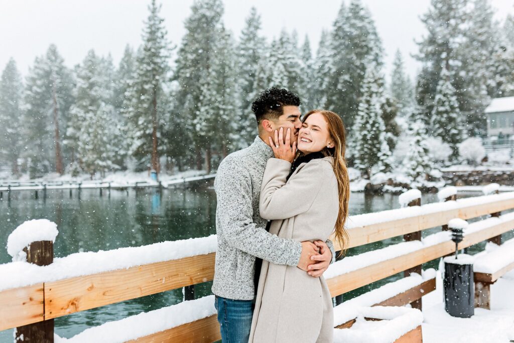 A couple stands on a snowy wooden dock by Lake Tahoe, embracing and smiling during a magical winter proposal, with snow-covered trees and cozy cabins in the background.