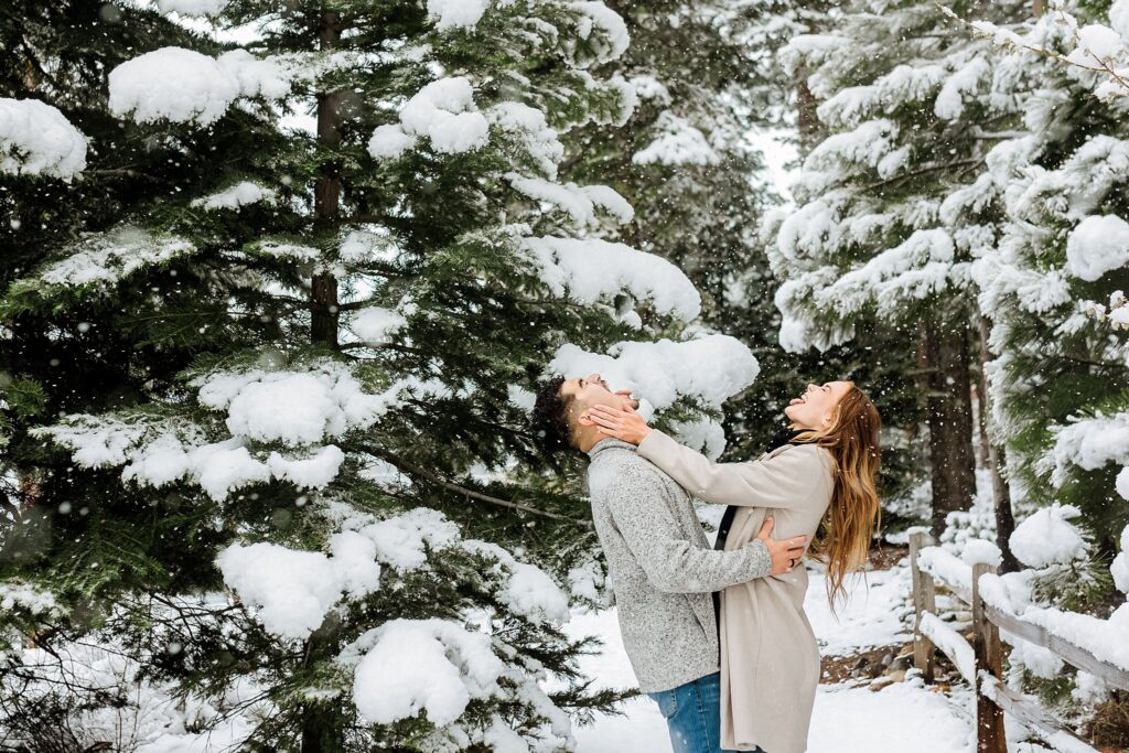 A woman playfully holds a man's face as they stand close together in a snowy forest near Lake Tahoe, both surrounded by snow-covered pine trees during a magical winter proposal.