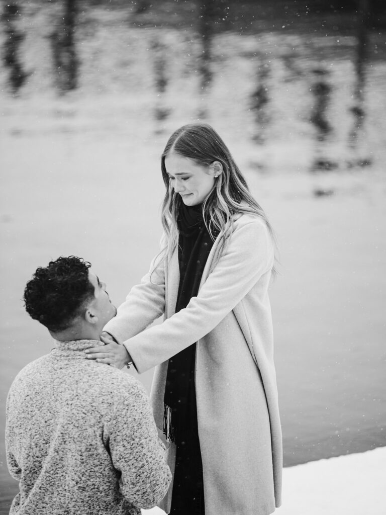 A man kneels in front of a woman by Lake Tahoe while snow falls; the woman is smiling and resting her hands on his shoulders, capturing a magical Lake Tahoe winter proposal.