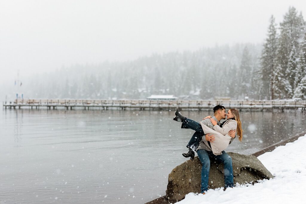 A couple sits on a large rock by a snowy lake, sharing a kiss as snow falls around them; the man holds the woman close in this Lake Tahoe Winter Proposal scene, with a dock extending over the icy water in the background.