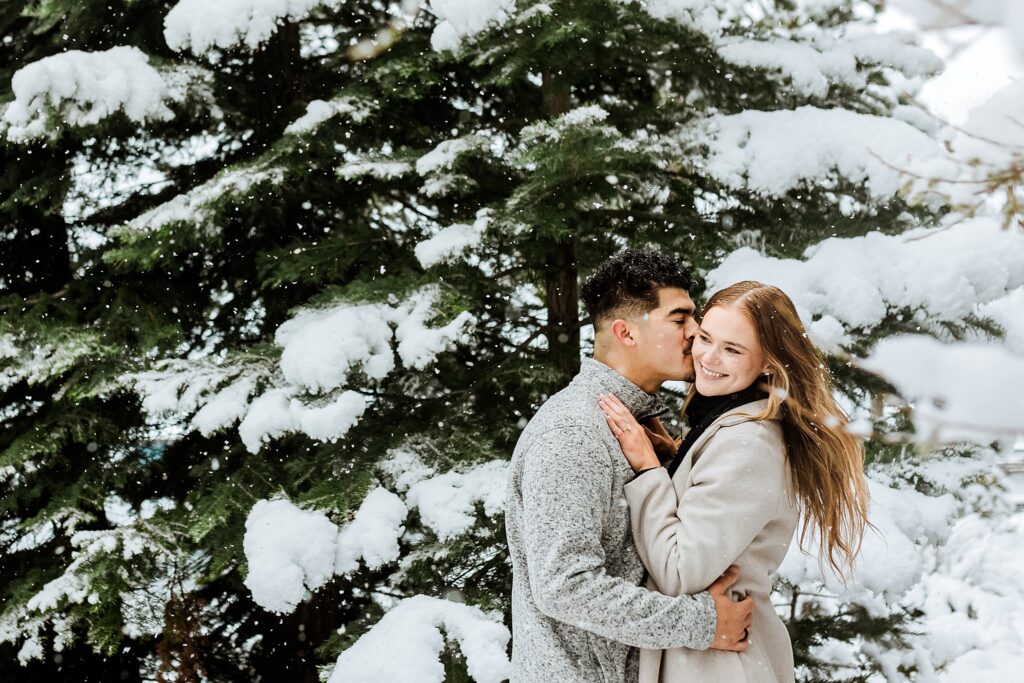 A couple embraces in front of a snow-covered evergreen tree during their Lake Tahoe Winter Proposal; the man kisses the woman’s cheek while she smiles at the camera.