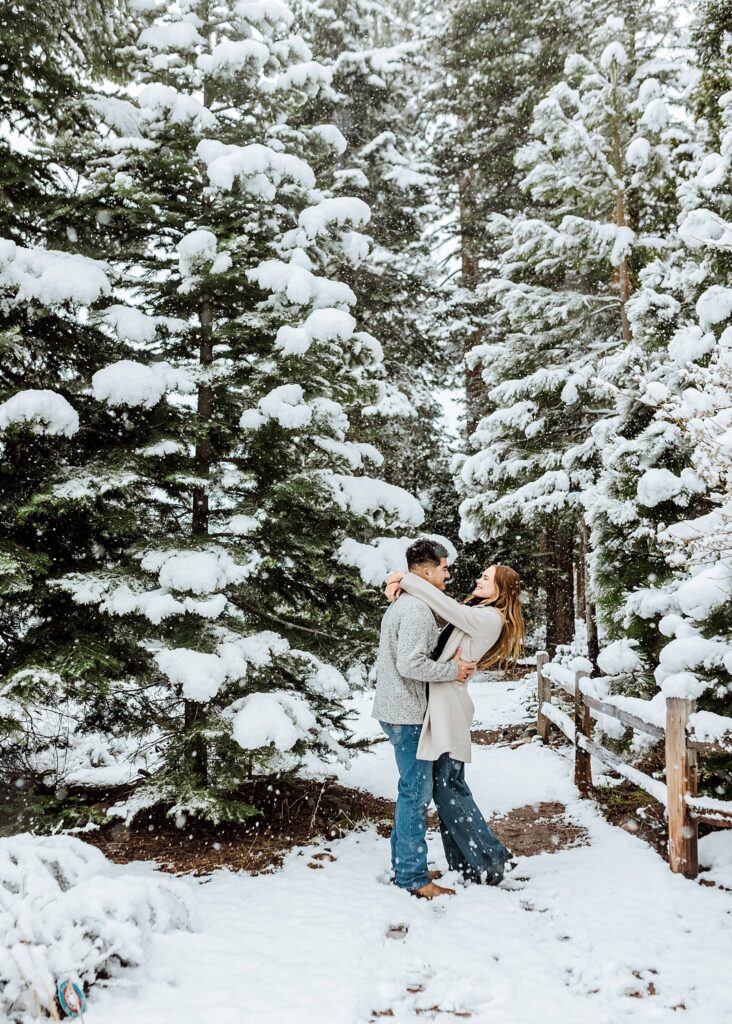 A couple stands embracing on a snow-covered path surrounded by tall, snow-laden evergreen trees in a forest setting.