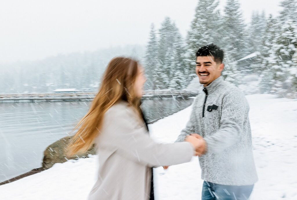 A man and a woman hold hands and smile while standing outdoors in falling snow near a lake with pine trees, capturing the magic of a Lake Tahoe winter proposal.