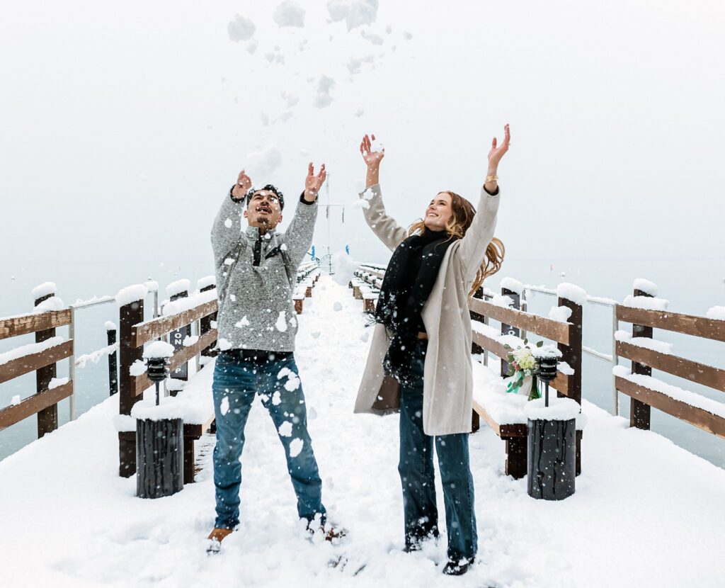 Two people stand on a snowy pier at Lake Tahoe, throwing snow into the air with their arms raised, surrounded by a magical winter landscape—perfect for a romantic winter proposal.