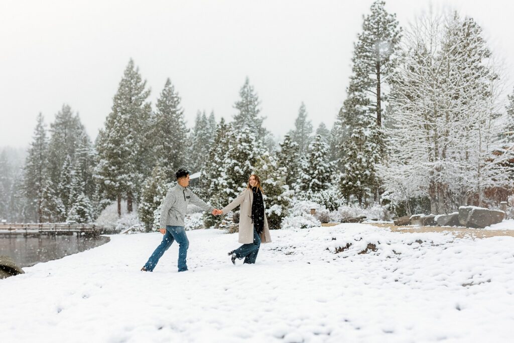 A couple holding hands walks through snow-covered ground near a lake, surrounded by snow-laden trees—a perfect Lake Tahoe Winter Proposal scene.
