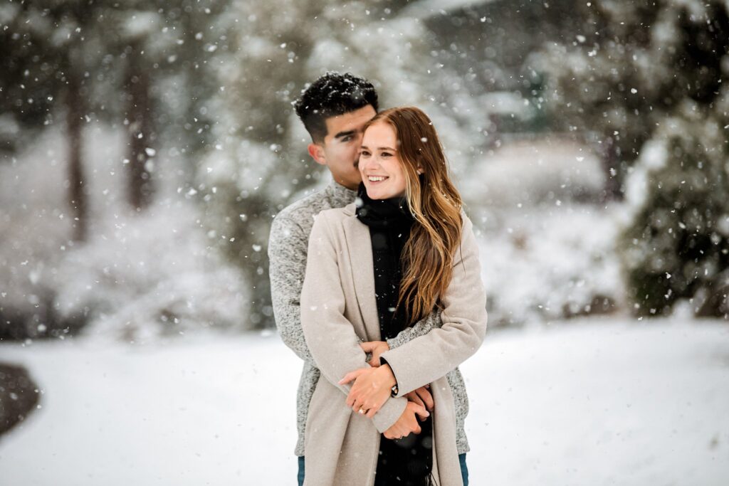 A man stands behind a woman, embracing her as they smile outdoors on a snowy day amid trees—a Lake Tahoe Winter Proposal to remember.