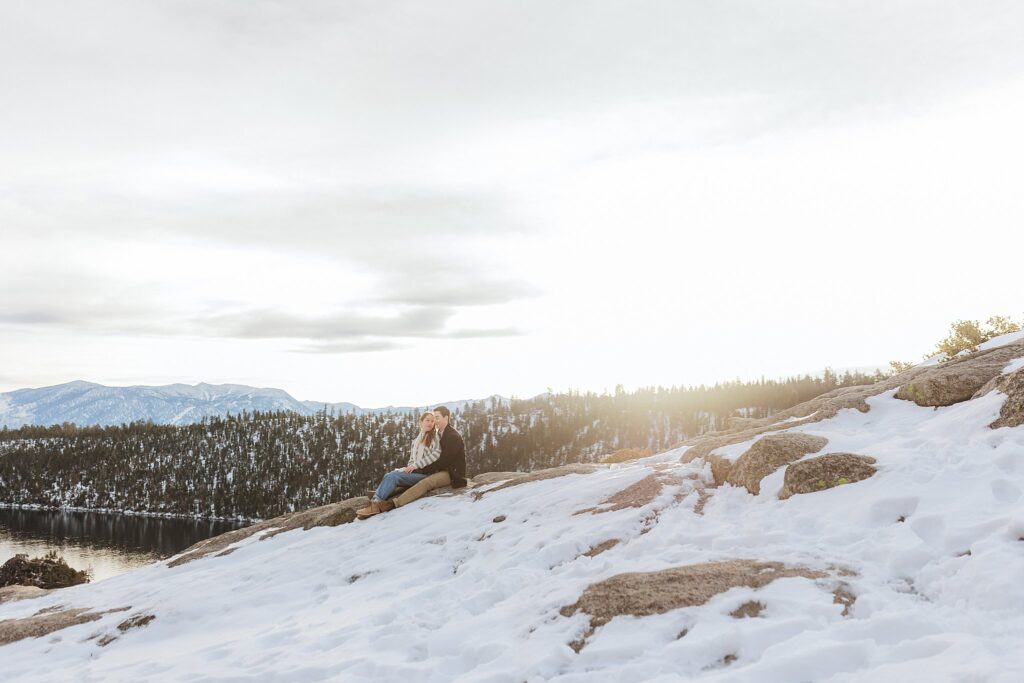 A person sits on a snow-covered rocky slope overlooking a forest and lake under a cloudy sky with sunlight in the background, capturing the serene beauty of a Lake Tahoe Winter Proposal.