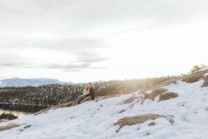Person sitting on a snowy, rocky hillside overlooking a forested lake and distant mountains under a cloudy sky, capturing the tranquil beauty of a Lake Tahoe winter proposal.