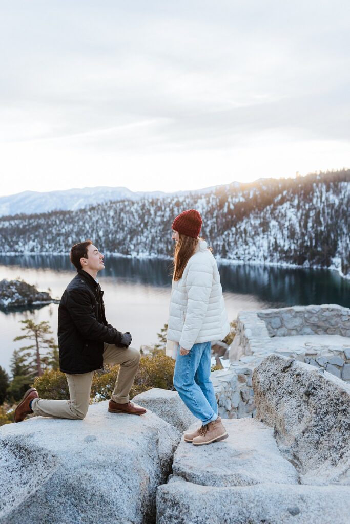 A man kneels on a rock proposing to a woman who is standing; they are outdoors near a lake with snowy mountains in the background, capturing the magic of a Lake Tahoe Winter Proposal.